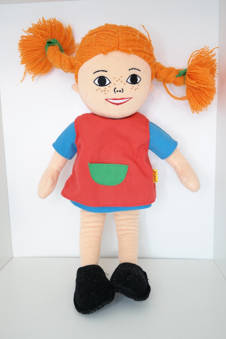 K&ouml;nnte beinhalten: Stoffpuppe mit orangefarbenen Z&ouml;pfen aus Garn, Sommersprossen und einem l&auml;chelnden Gesicht. Die Puppe tr&auml;gt ein rotes Kleid mit gr&uuml;ner Tasche und blauen &Auml;rmeln sowie schwarze Schuhe. Ein klassisches Kinderspielzeug.