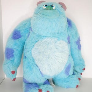 Puede incluir: Un peluche del personaje Sulley de la película Monstruos, S.A. El juguete es de color azul claro con manchas azules oscuras y una barriga blanca. Tiene orejas moradas, garras rojas y una expresión amigable.