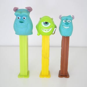 May include: Three plastic PEZ candy dispensers featuring characters from Monsters, Inc. The left is a blue Sulley with a light green stem, the center is a green Mike Wazowski with a yellow stem, and the right is a smaller blue Sulley with a brown stem.