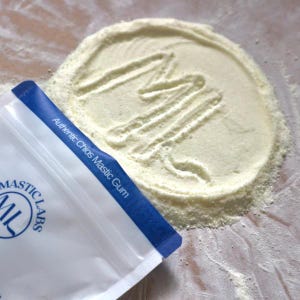 May include: A white bag with blue trim and the text "Authentic Chios Mastic Gum" and "Mastic Labs" printed on it. The bag is partially open and a white powder is spilling out, forming a circle with an "M" drawn in the powder.