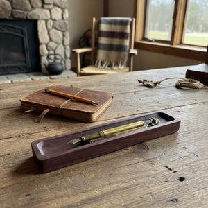 May include: A wooden desk with a leather-bound journal and pencil, a dark wood pen tray with a brass fountain pen, and a rustic interior setting with a fireplace and window.