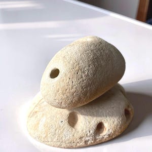 Op de afbeelding: Twee lichtbeige stenen, de ene rust op de andere. De bovenste steen heeft een enkel, groot gat. De onderste steen heeft verschillende kleinere gaten. De stenen hebben een ruw, getextureerd oppervlak, mogelijk strandstenen.