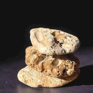 May include: Three beige, porous stones stacked. The stones have a rough texture and are lit by sunlight, creating shadows. The background is a dark, solid colour.