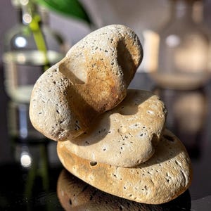 Op de afbeelding: Drie gladde, beige stenen op elkaar gestapeld, op een donkere, reflecterende ondergrond. De stenen hebben een natuurlijke, verweerde uitstraling met kleine gaatjes en kleurvariaties, variërend van lichtbeige tot warmbruin.