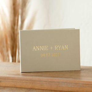 Puede incluir: Un libro de visitas de boda beige claro con letras doradas en relieve que dicen "Annie + Ryan 08.07.2023". El libro está sobre una mesa de madera con una taza blanca y un cuenco decorativo.