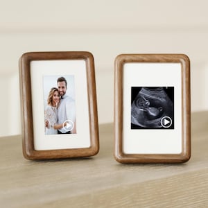 May include: Two wooden photo frames. One contains a photo of a couple, the other an ultrasound scan. The frames have a warm, brown finish and are displayed on a light-coloured surface.