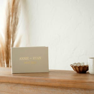 May include: A beige book with gold lettering that reads "ANNIE + RYAN" and a date. Beside the book is a small, brown bowl with white accents and a white mug. The items are on a wooden surface.