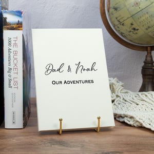 May include: A cream-colored journal with the text "Dad & Noah Our Adventures" in black script is displayed on a gold stand. Two books stand behind the journal, and a globe is in the background. The journal is a keepsake.