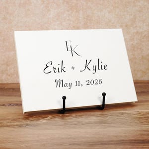 May include: A rectangular white sign displayed on a black metal stand. The sign features the black printed text EK at the top, followed by the names Erik + Kylie, and the date May 11, 2026, centered in a black serif font.