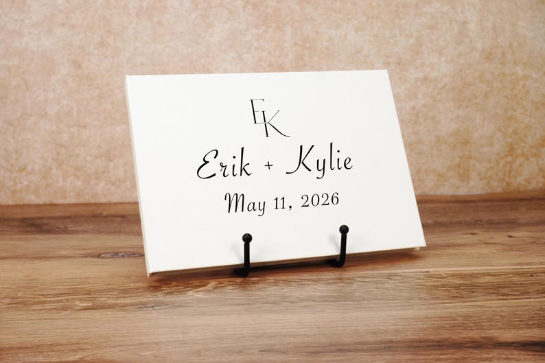 May include: A rectangular white sign displayed on a black metal stand. The sign features the black printed text EK at the top, followed by the names Erik + Kylie, and the date May 11, 2026, centered in a black serif font.
