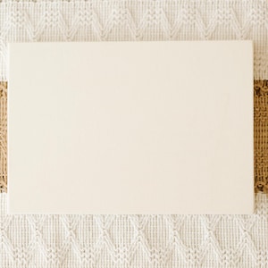 May include: A rectangular, smooth, off-white wooden board centered on a textured table runner featuring a cream-colored knit pattern with a wide, horizontal band of tan, woven, diamond-patterned fabric and a small white ceramic bowl.