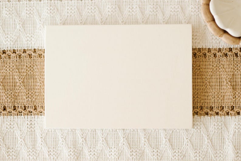 May include: A rectangular, smooth, off-white wooden board centered on a textured table runner featuring a cream-colored knit pattern with a wide, horizontal band of tan, woven, diamond-patterned fabric and a small white ceramic bowl.