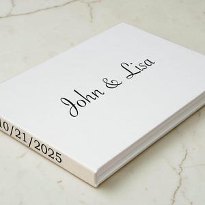 May include: A white rectangular guest book with a hard cover. The front cover features the black printed text John & Lisa in a cursive script font. The side spine of the book displays the black printed date 10/21/2025 in a simple serif font.