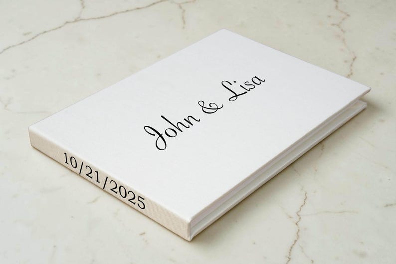 May include: A white rectangular guest book with a hard cover. The front cover features the black printed text John & Lisa in a cursive script font. The side spine of the book displays the black printed date 10/21/2025 in a simple serif font.