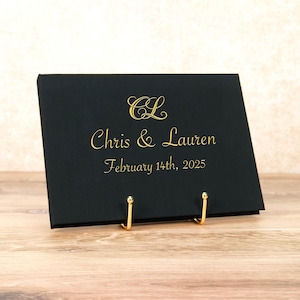 Könnte beinhalten: Ein schwarzes Hochzeitsschild mit goldfarbenen Buchstaben, auf dem "CL Chris & Lauren February 14th, 2025" steht. Das Schild wird von zwei goldfarbenen Metallständern gehalten. Der Hintergrund ist eine hellbeige Wand und ein Holztisch.