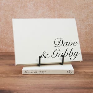 May include: A rectangular white canvas sign featuring the black printed text Dave & Gabby, supported by a black metal stand. A smaller white rectangular base sits underneath, displaying the black printed text March 18, 2026 and two hearts.