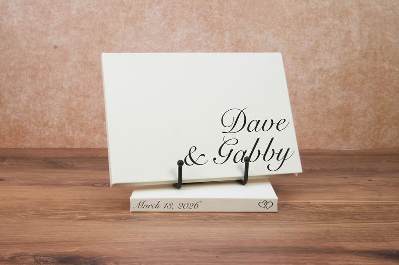 May include: A rectangular white canvas sign featuring the black printed text Dave & Gabby, supported by a black metal stand. A smaller white rectangular base sits underneath, displaying the black printed text March 18, 2026 and two hearts.