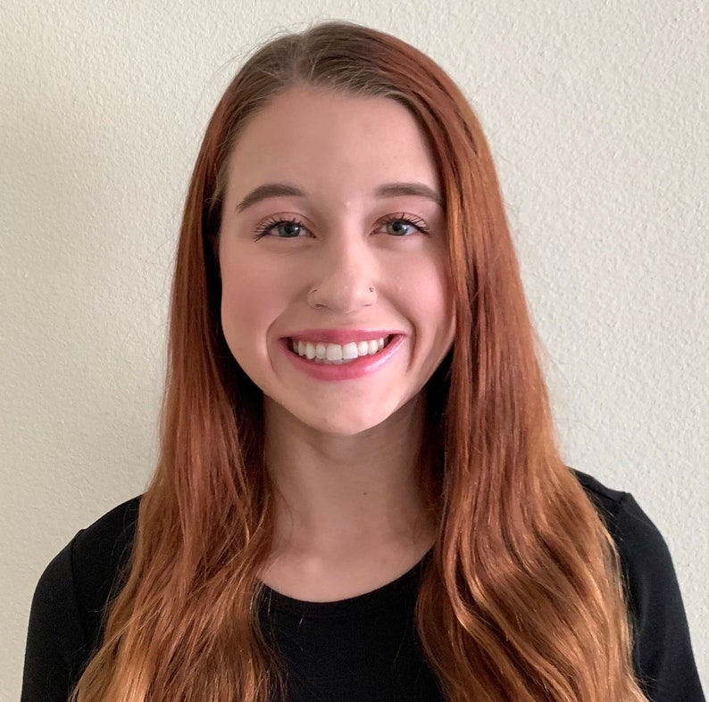 May include: A woman with long, wavy, auburn hair is smiling at the camera. She is wearing a black, long-sleeved shirt.