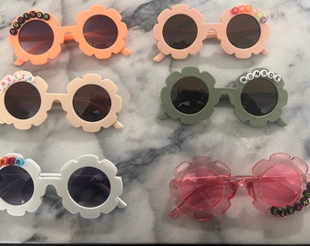 May include: Five pairs of colorful flower-shaped sunglasses with beaded names, including Addison, Tressa, Monroe, Parker, and Eleanor.