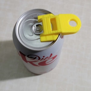 Könnte beinhalten: Gelber Plastik-Dosenöffner, der oben auf einer silbernen Dose befestigt ist. Die Dose ist mit "Diet Coke" beschriftet.