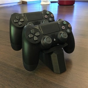 May include: Two black wireless game controllers are mounted on a black charging stand. The controllers have buttons and joysticks.