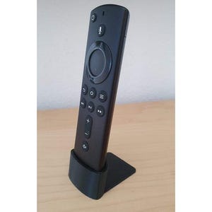 May include: A black remote control with a circular button in the center and a variety of buttons on the sides. The remote is sitting on a black stand.