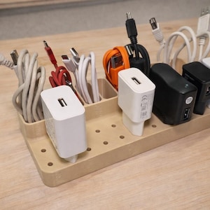 Puede incluir: Una estación de carga de madera con múltiples compartimentos que contienen varios cables de carga y adaptadores de corriente. Cables en blanco, rojo y naranja. Adaptadores de corriente blancos y negros.
