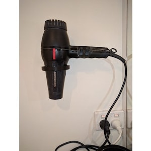 Puede incluir: Un secador de pelo negro con un acento rojo en el mango. El secador está montado en una pared blanca con una toma de corriente blanca debajo. El secador tiene el texto "Travel Dryer 2000" impreso en el lateral.