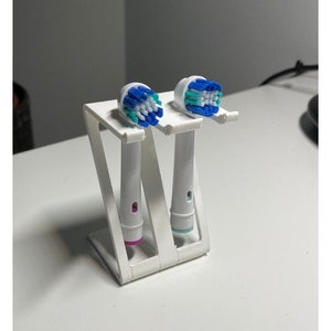 May include: A white toothbrush holder with two electric toothbrush heads. The toothbrush heads have blue and white bristles. The holder is made of white plastic and has a modern, minimalist design. The holder is designed to hold two toothbrushes.