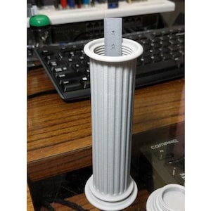 Puede incluir: Un recipiente blanco en forma de columna impreso en 3D con un diseño acanalado. El recipiente está vacío y tiene una regla en su interior.