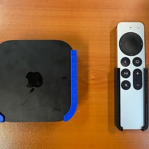 Puede incluir: Un dispositivo Apple TV negro con un soporte azul y un mando a distancia plateado con un soporte negro. El Apple TV tiene el logotipo de Apple.