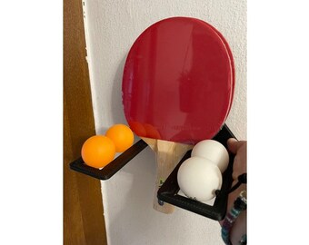 May include: A black wall-mounted rack with two ping pong paddles and three orange and white ping pong balls.