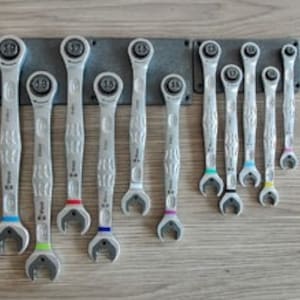 Könnte beinhalten: Ein Satz silberner Ratschenschlüssel in verschiedenen Größen, ordentlich auf einem dunkelgrauen Metallhalter angeordnet. Jeder Schlüssel hat ein farbiges Band in der Nähe des Fußes, und die Größen sind oben markiert.