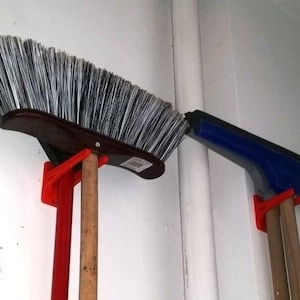 May include: Two brooms and a squeegee are hanging on a wall using red plastic wall mounts. The brooms have wooden handles and the squeegee has a blue handle.