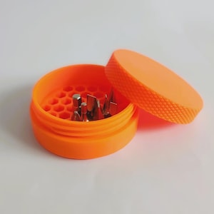 May include: Bright orange round container with a matching lid, partially open to reveal various metal screwdriver bits. The container has a textured lid and a honeycomb-patterned interior. The items are set against a white background.