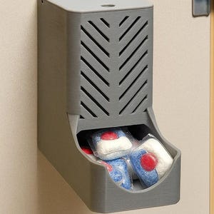 May include: A gray, wall-mounted dispenser with a chevron-patterned vent, holding several blue and white dishwasher detergent pods with red accents. The dispenser has a small opening at the top and a curved opening at the bottom for easy access.