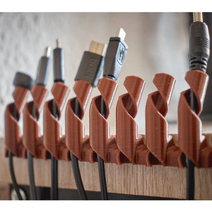 Puede incluir: Un conjunto de organizadores de cables de color marrón hechos de plástico flexible, diseñados para sujetar y organizar cables y alambres. Los organizadores están retorcidos en forma de espiral y están unidos a una superficie de madera.
