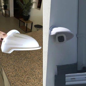 Puede incluir: Cubierta de cámara de seguridad de plástico blanco con parte superior redondeada y fondo plano. La cubierta está diseñada para proteger la cámara de los elementos y disuadir el vandalismo.