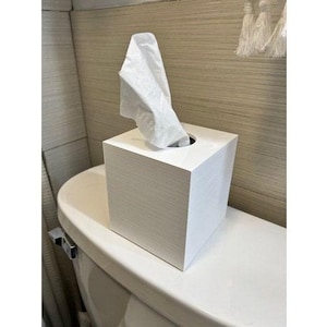 Puede incluir: Un dispensador de pañuelos cuadrado blanco con un pañuelo blanco sacado. El dispensador de pañuelos está sobre una encimera blanca.