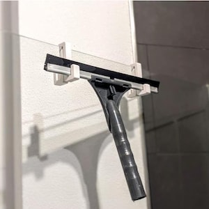 Puede incluir: Un soporte de escobilla de goma de plástico blanco con una escobilla de goma negra unida a él. La escobilla de goma está colgada en una puerta de ducha de vidrio.