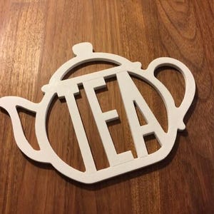 May include: A white wooden teapot-shaped sign with the word "TEA" cut out in the centre. The sign is placed on a wooden surface. The teapot design includes a handle, spout, and lid.