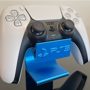 以下が含まれることがあります： 白い PlayStation 5 コントローラーが、PlayStation ロゴの付いた青いスタンドに置かれています。スタンドはプラスチック製で、表面が滑らかです。