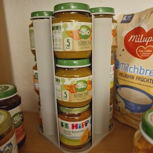 Może przedstawiać: Prezentacja słoiczków z jedzeniem dla niemowląt, głównie puree z dyni, z zielonymi wieczkami. Słoiczki są ułożone w obrotowym stojaku ekspozycyjnym. W tle widoczna jest torebka z kaszką dla niemowląt.