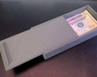 Puede incluir: Una caja rectangular gris con una tapa corredera, parcialmente abierta para revelar una pila de billetes de veinte dólares. La caja está hecha de un material sólido y mate y se encuentra sobre una superficie oscura.