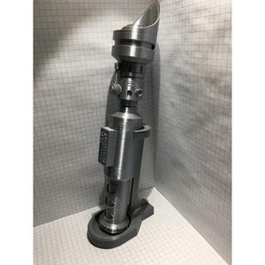 以下が含まれることがあります： グレーの3Dプリントされたスターウォーズライトセーバーの小道具のモデル。ライトセーバーは、テクスチャ加工された表面と丸みを帯びたトップを持つ円筒形のボディを持っています。ライトセーバーはグレーのベースに取り付けられています。