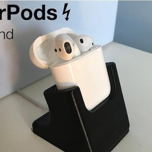 Puede incluir: Un soporte negro con una funda de carga AirPods blanca. El soporte tiene un recorte para la funda y el texto "AirPods ⚡️ Stand" es visible en el fondo.