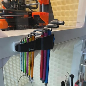 May include: A set of multi-coloured hex keys, arranged in a black holder. The keys are in various colours, including yellow, green, blue, and red. The holder is mounted on a white surface, with the brand name "Wera" visible.