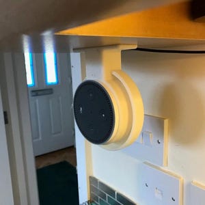 Peut inclure: Un support d'enceinte intelligent imprimé en 3D de couleur blanche, fixé sous un meuble. L'enceinte est noire et ronde, avec un microphone et des boutons sur le devant.