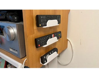 May include: Three white plastic remote control holders mounted on a wooden wall. Each holder has a curved design and holds a black remote control.