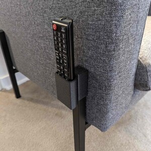 Puede incluir: Un mando a distancia negro se encuentra en un soporte negro impreso en 3D que está unido al lado de una silla gris.